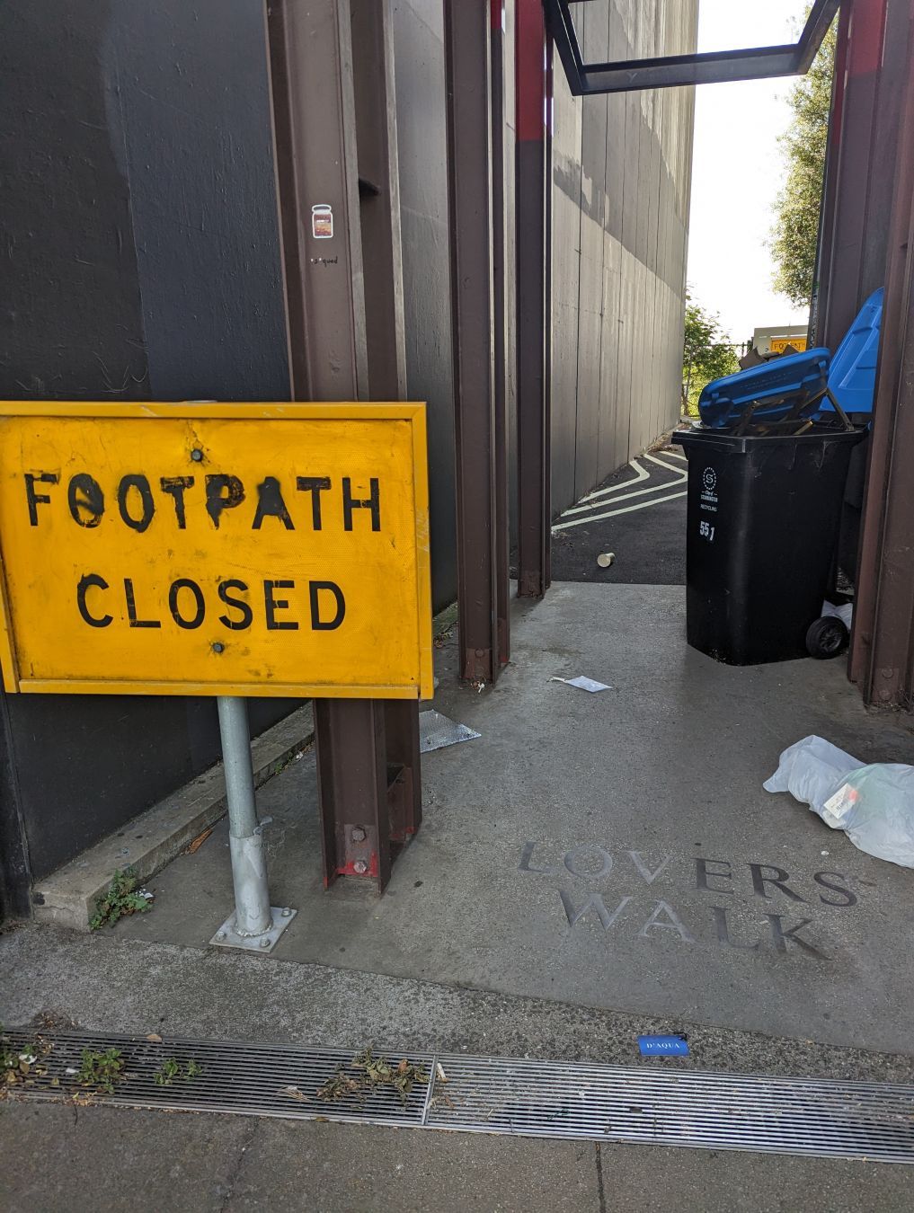 An grey laneway with Lovers Walk embossed at the start of it. A yellow sign reads Footpath closed.

The laneway is empty with the exception of white garbage bags and large green garbage bins.