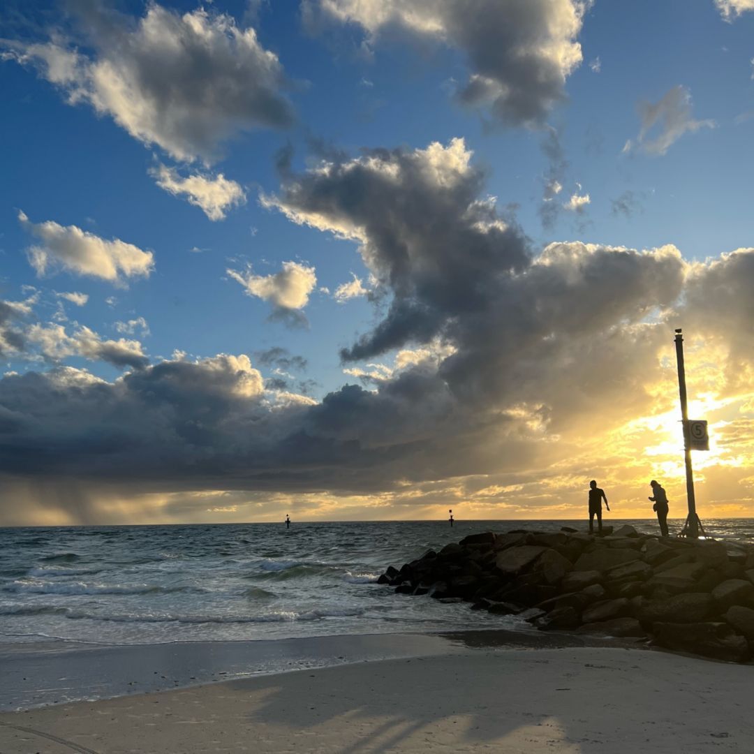 A cloudy sunset over a calm beach with a rock wall in the foreground. Two silhouetted figures are standing on the wall.