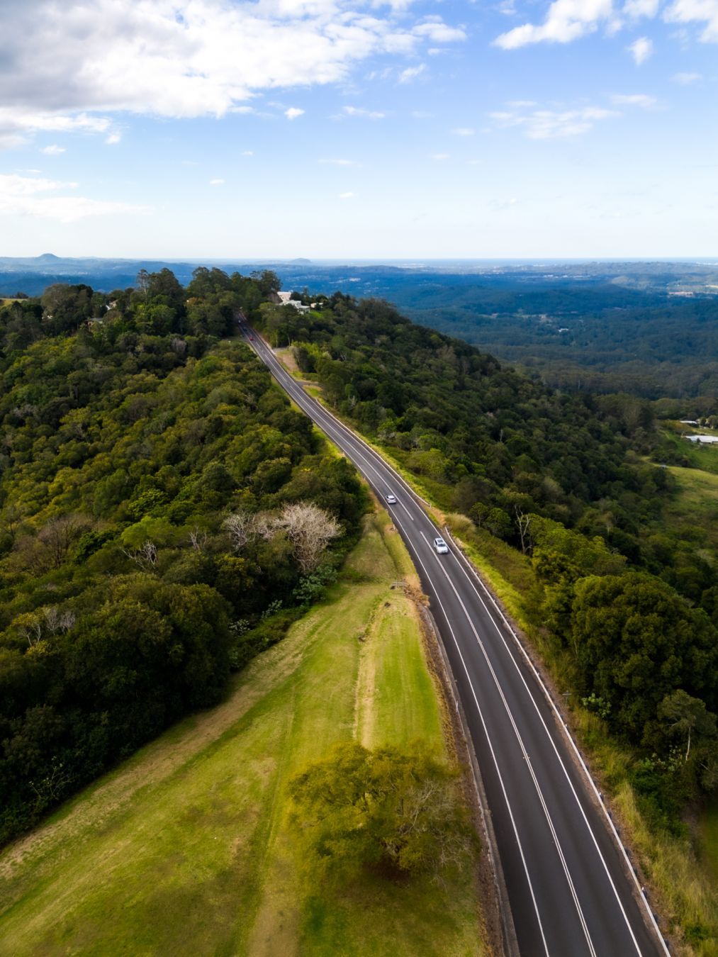 Overhead photo of a two lane road on top of a hill with hinterland and coastline in view in the distance.