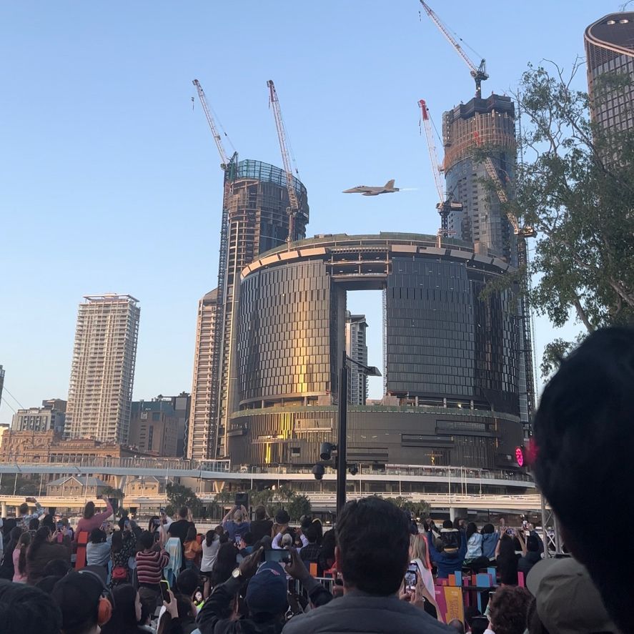 A Boeing EA-18G Growler jet aircraft flies from right to left, over the Brisbane river, in front of several Brisbane high-rises including the casino currently under construction. Many people are watching.