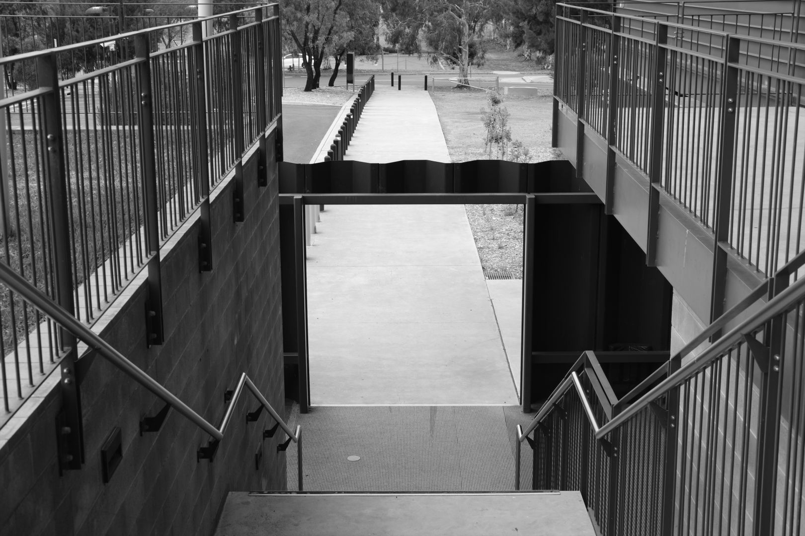 A black and white photo looking down a flight of concrete steps and through an archway in a metal fence. Metal fences around the stairway give a strong vertical rhythm and a slight feeling of entrapment and escape