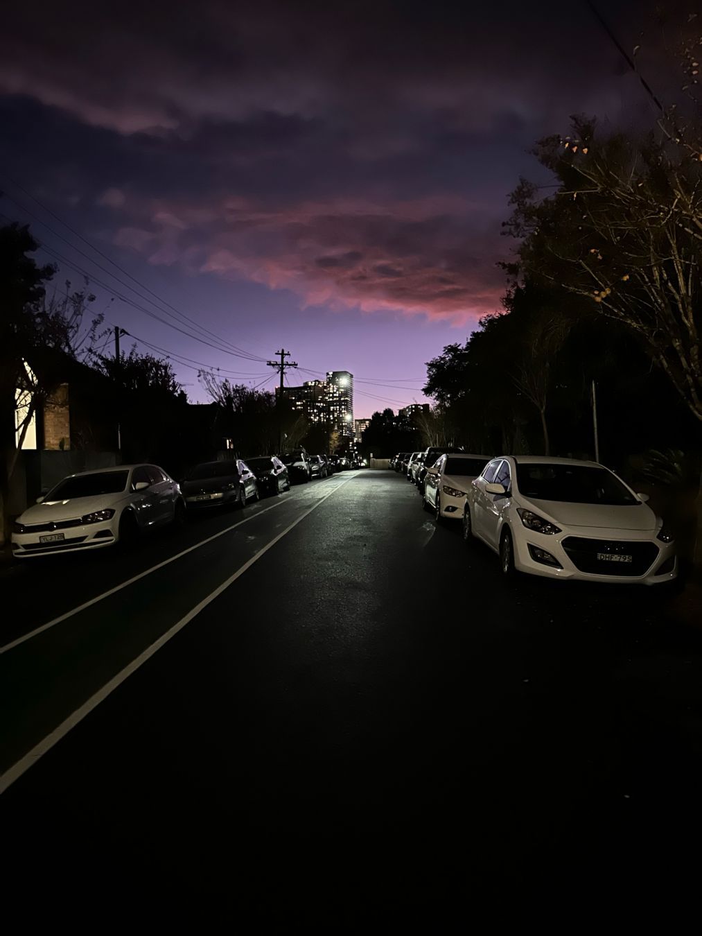 A dark street lined with cars, below a pinkish dusk sky, a street light reflects off the wet road surface