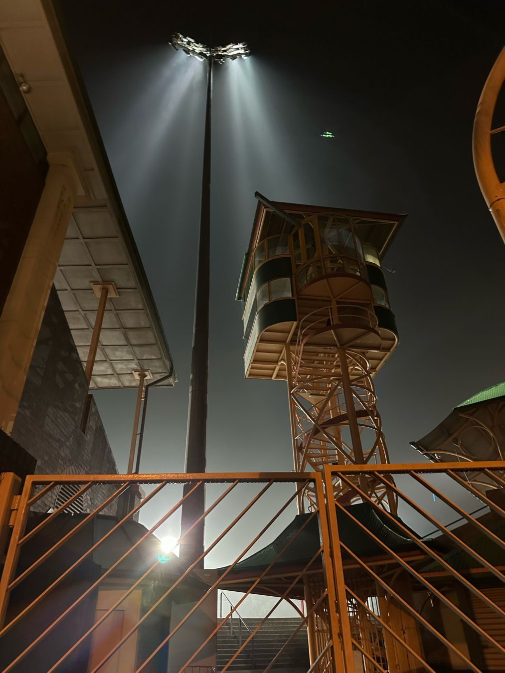 Looking up at the commentators’ tower and a light tower, North Sydney oval at night. The lights are on, lighting the misty air